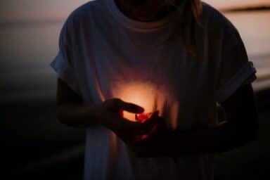 Person in a white t-shirt cupping a warm glowing light in their hands against a dark, coastal background at dusk.