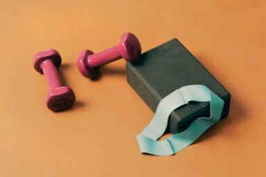 Two pink dumbbells, a black yoga block, and a light blue resistance band rest on an orange surface, representing limited mobility fitness.