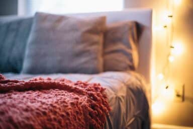 Fuzzy pink blanket on a bed with grey pillows and warm string lights glowing in the background.