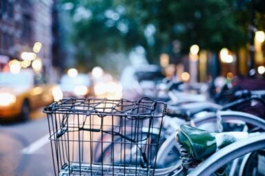 A row of parked bicycles with a prominent black metal basket and a green seat, set against a blurred city background with glowing vehicle headlights and streetlights at dusk.