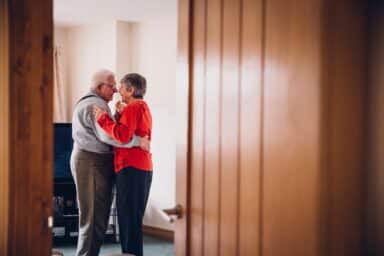 An elderly couple dances closely together in a living room, framed by a wooden doorway.