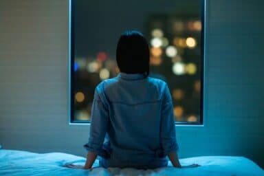 A woman in a denim shirt sits on a bed, looking out a window at city lights during the night.