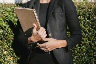A person in a professional black blazer and dress holds a tan folder while standing in front of a green hedge.