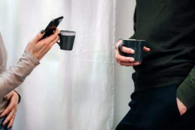 Two people stand near a white curtain holding coffee mugs, with one person checking a smartphone during the conversation.