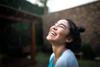 A woman with dark hair in a bun tilts her head back and laughs heartily outdoors, representing laughter is the best medicine.
