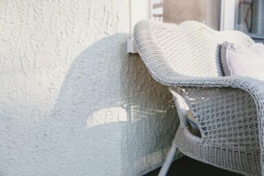 A white wicker loveseat with a soft pillow sits against a textured white wall in bright sunlight.