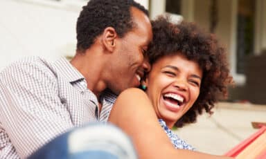 A man whispers into a laughing woman's ear as they sit together outdoors in a hammock.