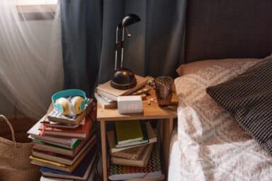 A digital alarm clock displaying 11:56 sits on a wooden nightstand cluttered with books, a lamp, and headphones next to a bed.