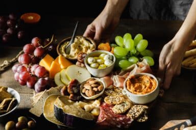 A person's hands place a wooden platter filled with grapes, cheese, nuts, hummus, olives, and sliced meats on a dark table, representing the Mediterranean diet.