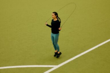 A woman with a long dark braid jumps rope on a green outdoor court with white lane lines, representing the mental health benefits of exercise.