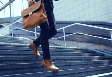 A person dressed in business attire is shown walking down a set of outdoor stone stairs while carrying a brown leather briefcase.