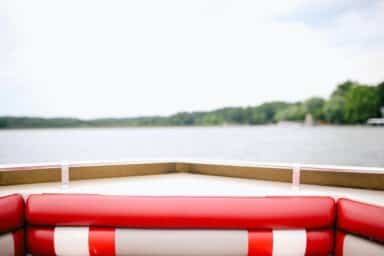 A view from the front of a boat with red-and-white cushioned seats looks out over a calm lake toward a distant, tree-lined shore.