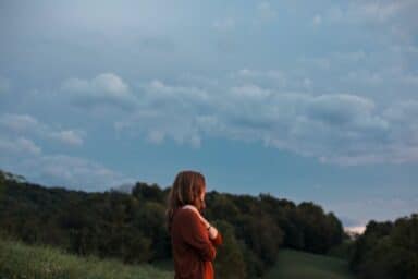 A person with long brown hair stands in a grassy field at dusk, looking out over a rolling forest under a cloudy blue sky.