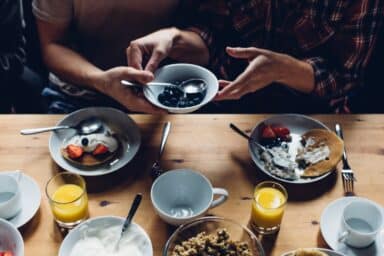 Two people share a bowl of blueberries over a healthy breakfast of pancakes, fruit, and orange juice.