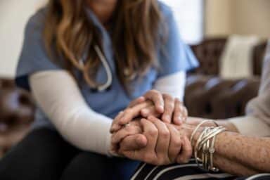 A nurse in blue scrubs gently holds the hands of an elderly patient in a supportive gesture.