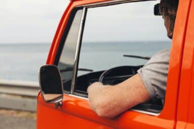 A man rests his freckled arm out of the window of an orange van as he drives alongside the ocean, representing the freedom found in overcoming drug addiction.