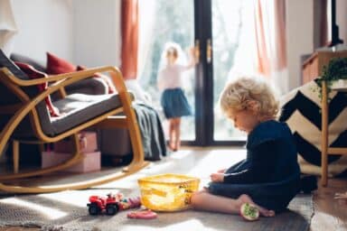 An image shows a young girl in a blue dress sitting on a rug and playing with toys, while another child stands in the background looking out of a glass door.