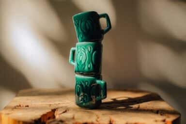 Three small, teal ceramic mugs stacked vertically on a rough-hewn wooden block, casting a shadow against a neutrally toned, dappled wall.