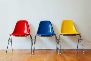 Red, blue, and yellow molded plastic chairs stand in a row against a white wall on a wooden floor.