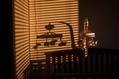 A mobile hanging over a crib casts long, dark shadows against a wall through window blinds, representing postpartum depression.