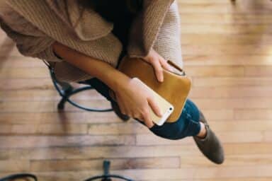 A person sits on a stool with a smartphone in their lap, resting atop a brown leather wallet or clutch.