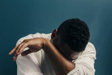 A Black man hides his face in his arm, leaning forward in a state of distress against a dark blue background.
