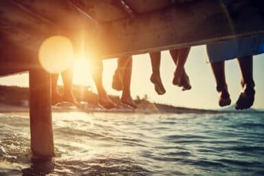 Four pairs of legs and feet dangled from a wooden pier over the ocean during a golden sunset.
