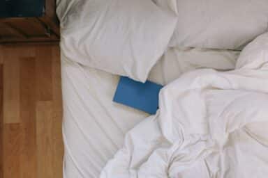 A blue hardcover book resting on unmade white bed sheets and pillows next to a wooden nightstand.