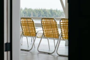 Three yellow and white woven lawn chairs sit empty on a deck overlooking a forest and water.