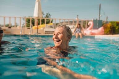 A senior woman with short gray hair smiles joyfully while swimming in a bright blue outdoor pool, representing fitness as you age.