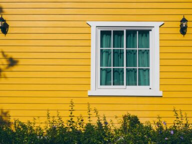 A white window frame with green curtains set against a bright yellow house exterior with small lanterns, representing senior housing options.