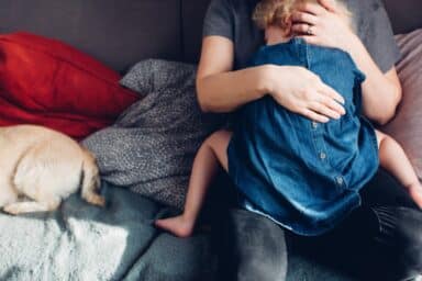 A woman in a grey shirt sits on a sofa and tightly embraces a young child in a blue denim dress, representing separation anxiety.