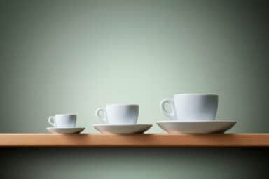 Three white espresso and coffee cups with saucers in small, medium, and large sizes on a wooden shelf.