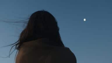 Silhouette of a person with long hair looking at a small, bright moon in a dark blue twilight sky.
