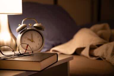 Eyeglasses rest on a closed book next to an alarm clock on a bedside table under a warm lamp.