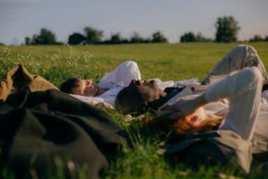 Three people lie together in a grassy field, laughing and talking under a clear sky.