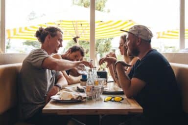 Four people sit at an indoor table talking over a meal, with yellow-and-white striped umbrellas visible through the window behind them.