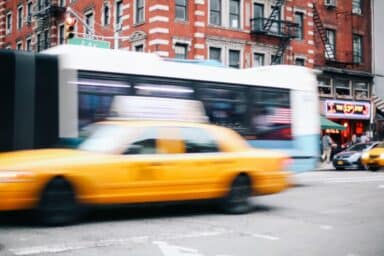 A yellow taxi and city bus blur as they speed through a busy New York City street corner.