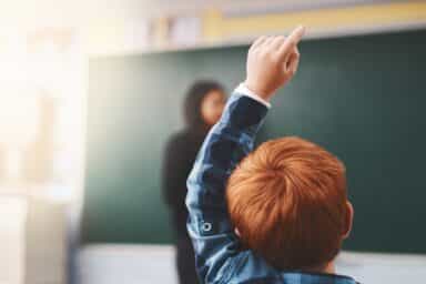 Redheaded student raising their hand in a classroom while a teacher stands at the chalkboard, illustrating teaching students with ADHD.