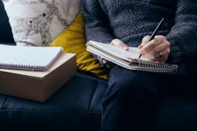A person in a charcoal knit sweater sits on a dark sofa, writing in a spiral notebook, representing therapy for anxiety disorders.