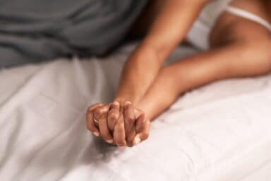 A couple holds hands with interlaced fingers on a bed, illustrating physical intimacy and emotional connection.