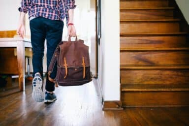 Person carrying a brown backpack walks toward an open doorway past a wooden staircase, symbolizing the path of treatment for adult ADHD.