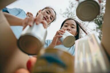 Two women look down into a cardboard box while packing canned food donations, representing volunteering.