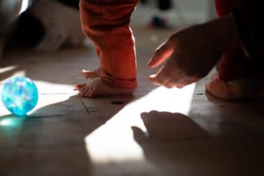 A toddler's bare feet on a wooden floor near a caregiver's hand, illustrating secure attachment and bonding.