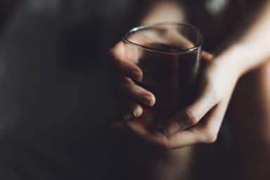 A person's hands are shown tightly gripping a small glass filled with a dark liquid in a dimly lit setting.
