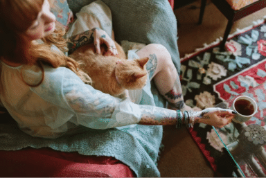 Person with tattoos on their arms sits in a chair, petting an orange cat on their lap while reaching for a white mug of tea.