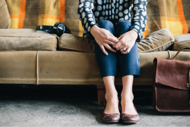 Person sits on a tan couch with hands clasped over knees, a leather bag on the floor, and headphones nearby.