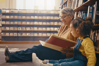 Older adult and young child sitting on a library floor, reading a book together in front of bookshelves filled with books.