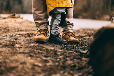Child in a yellow jacket standing between an adult's feet in a natural, outdoor setting with a dirt ground.