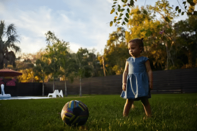 Young child in a blue dress stands on a green lawn looking toward a colorful soccer ball, with trees and a fence in the background.